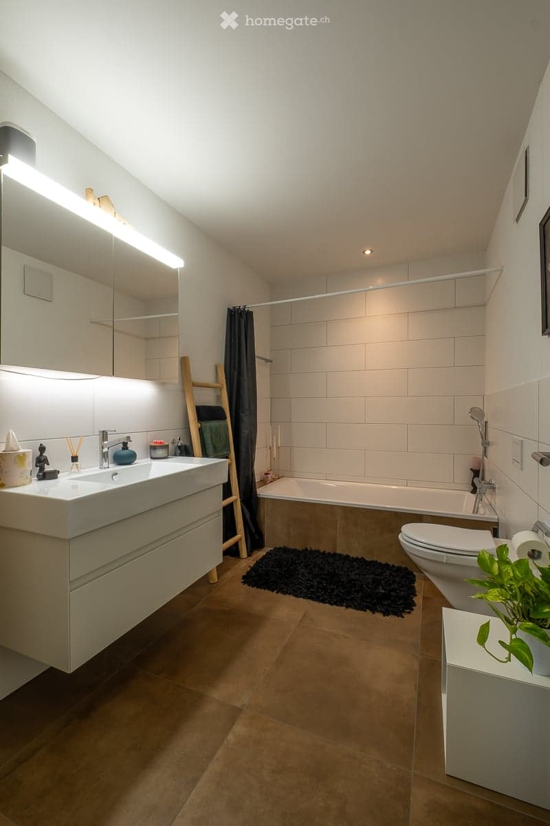 Modern bathroom with a large sink, mirror, bathtub, toilet, and a small plant on a white shelf. The room features wooden flooring, white tiles, and a black rug in front of the bathtub.