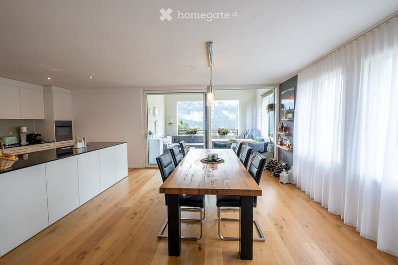 Open-plan kitchen and dining area with wooden flooring, white cabinets, a large wooden dining table with black chairs, and a view of a balcony with outdoor seating.
