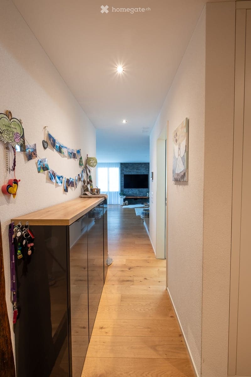 Bright hallway with wooden flooring, recessed ceiling lights, and a small wooden console table with hooks for keys and leashes. Photos and decorations are displayed on the walls.