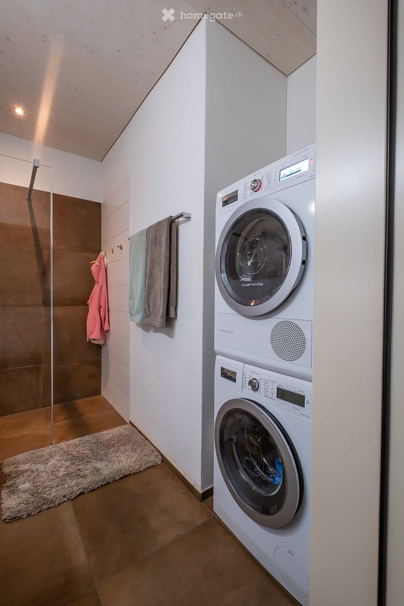 Modern bathroom with a stacked washer and dryer, tiled walls, wooden floor, and a small rug.