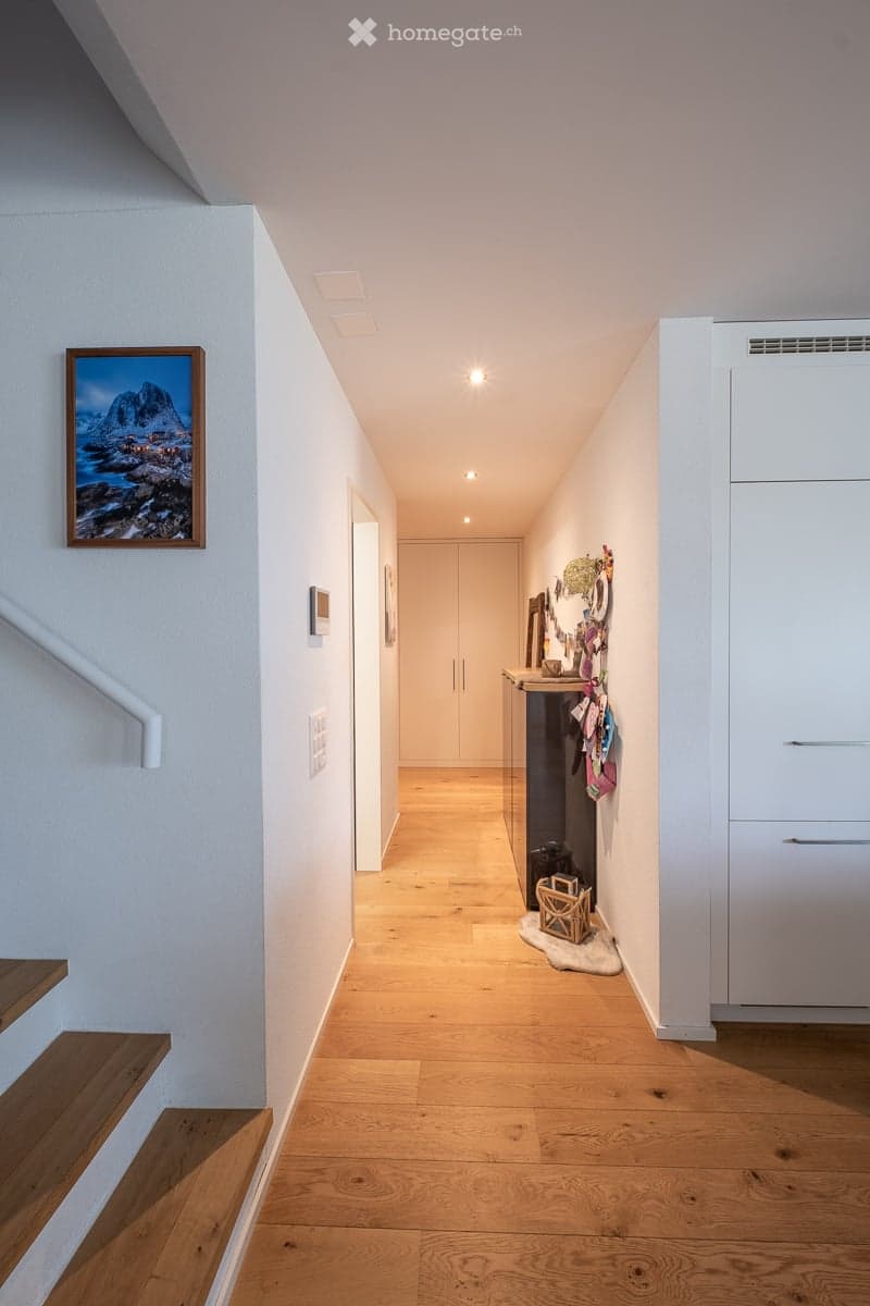 A modern hallway with light wooden flooring, white walls, and recessed ceiling lighting. Features include a staircase on the left, a small console table with a decorative item, and a framed picture on the wall.