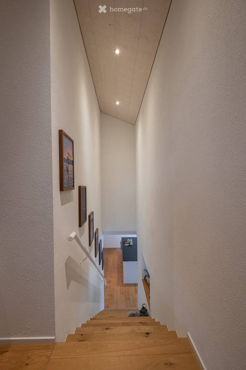 A narrow, modern hallway with wooden stairs, white walls, and framed artwork. The ceiling has recessed lighting, and the floor is wooden.