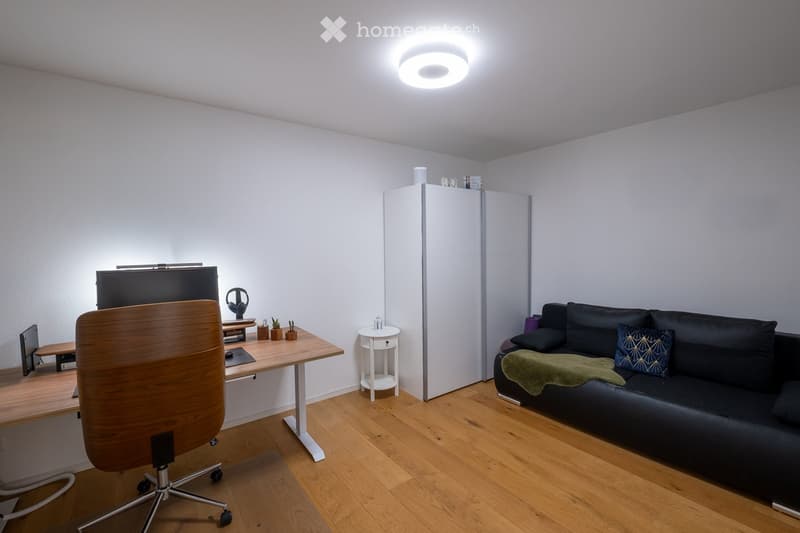 A modern, well-lit room with a black sofa, a wooden desk with a chair, a screen, and a small white table with a cup. The room features a wooden floor and white walls.