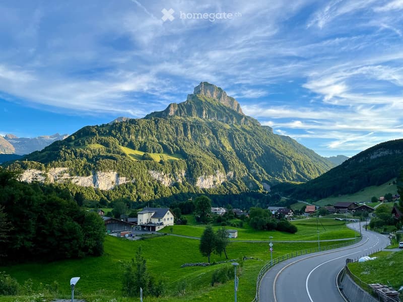 A picturesque mountain valley with a winding road, scattered houses, and lush green meadows surrounded by towering peaks.