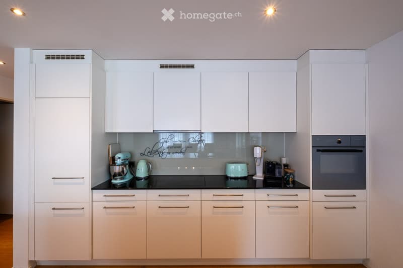 Modern kitchen with white cabinets, stainless steel handles, and a black countertop. Appliances include a refrigerator, oven, mixer, toaster, and coffee maker.