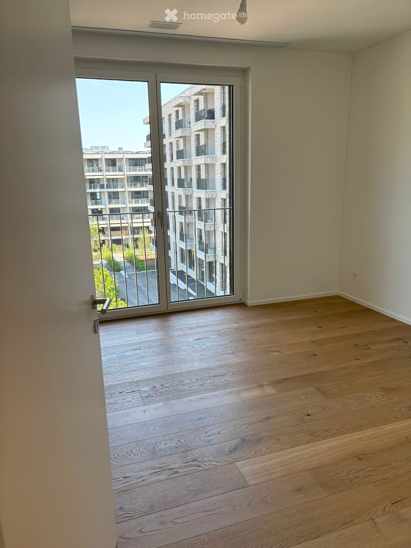 Empty room with wooden flooring and large sliding glass doors leading to a balcony with a view of a modern apartment building.