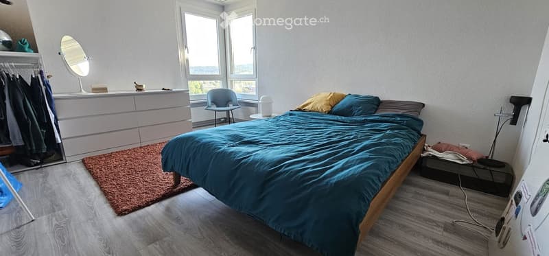 Cozy bedroom with a wooden bed, blue duvet, a small chair, a mirror, a dresser, and a clothing rack. The room has large windows with a scenic view and a warm, inviting atmosphere.