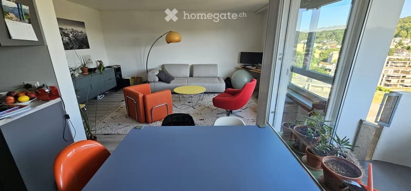 Modern living room with large windows offering a scenic view, featuring a grey sofa, orange armchairs, a yellow coffee table, a floor lamp, and a screen on a desk.