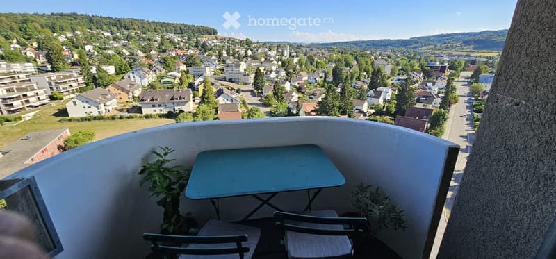 Balcony with a table, chairs, and plants, offering a panoramic view of a town and surrounding hills.