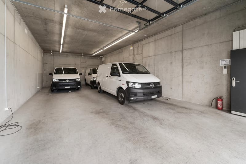 Underground garage with three white vans parked inside, concrete walls and ceiling, fluorescent lighting, and a fire extinguisher near the door.