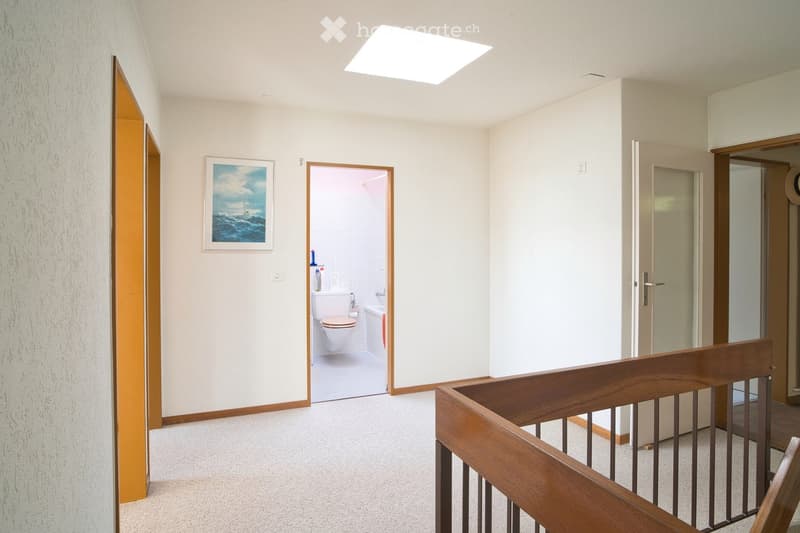 A well-lit hallway with carpeted flooring, white walls, and a wooden staircase railing. A painting hangs on the wall, and an open door leads to a small bathroom with a toilet, bathtub, and various toiletries.