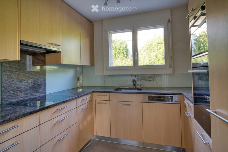 Modern kitchen with light wooden cabinets, stainless steel appliances, granite countertop, and a large window overlooking greenery.