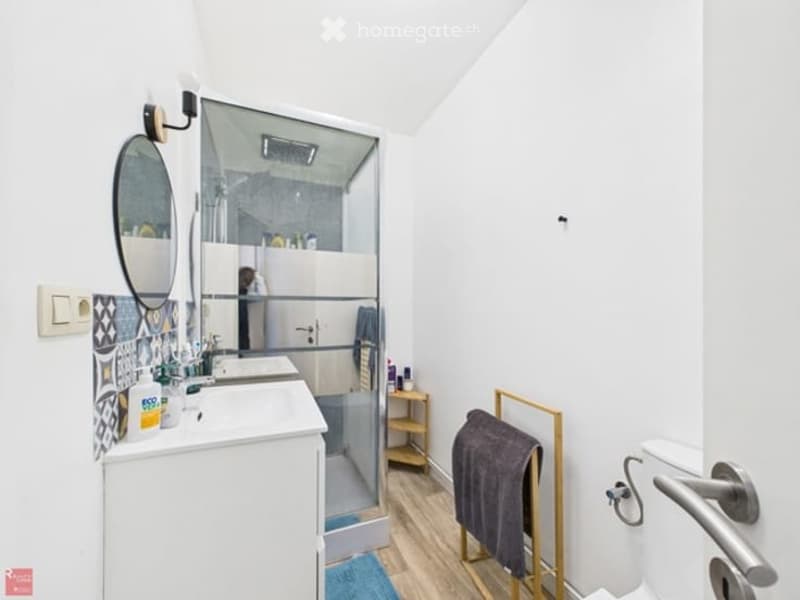 Modern bathroom with white walls, wooden flooring, a large mirror, a sink with a cabinet, a shower with a glass enclosure, a towel rack with a towel, and a small shelf holding toiletries.