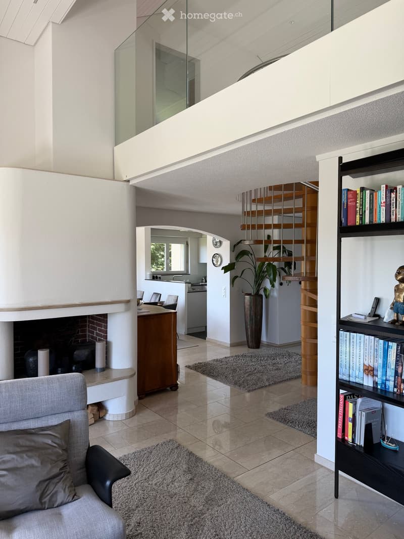 Modern living room with a grey sofa, bookshelf, fireplace, and a small kitchen area visible in the background.