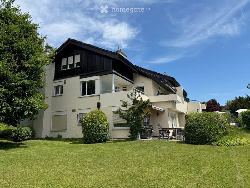 Two-story house with a modern design, featuring a balcony, large windows, and a well-maintained garden with a patio and outdoor furniture.