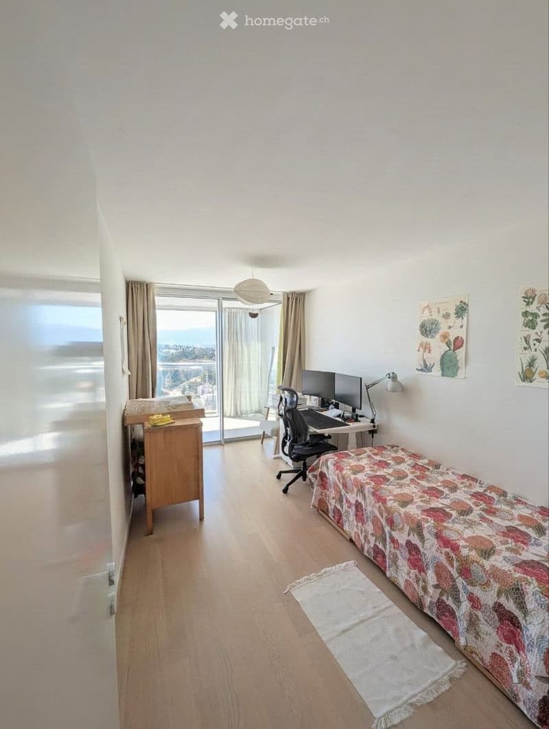 Bright room with wooden flooring, large window with view, desk with computer, chair, bed with floral cover, rug, and cabinet.