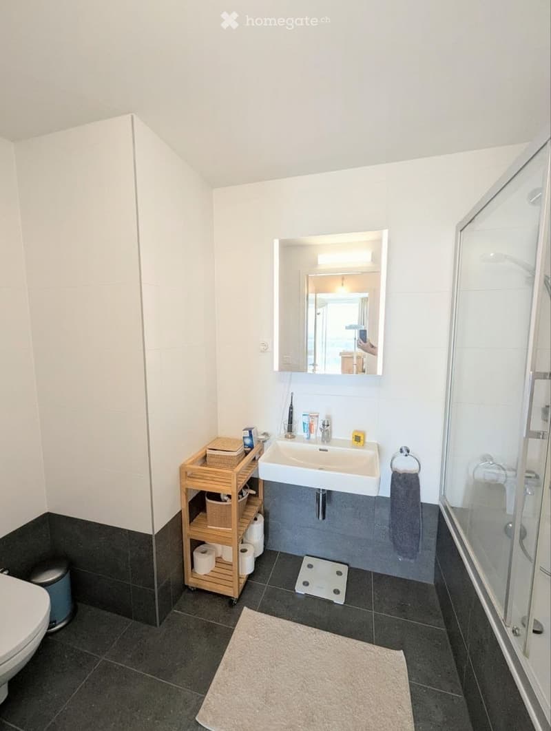 Modern bathroom with a white sink, mirror, toilet, wooden shelf, and a glass-enclosed shower. The floor is tiled in dark gray, and the walls are white with a black lower half.