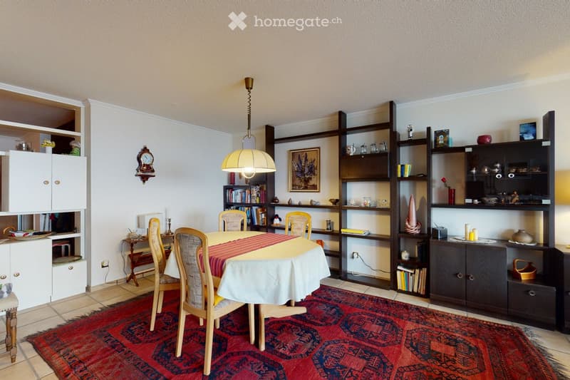 Dining room with a round table covered in a white tablecloth and red runner, surrounded by four wooden chairs. A large bookshelf filled with books and decorative items is visible, along with a wall-mounted clock and a pendant light hanging above the table
