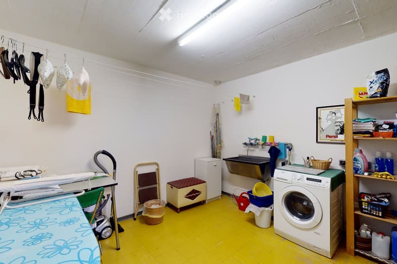 Brightly colored bathroom with yellow flooring, white walls, and various cleaning supplies. Includes a washing machine, ironing board, shelves, sink, and a small stool.