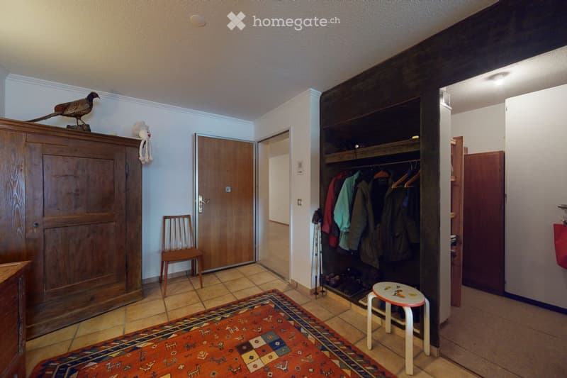 The image shows a hallway with a wooden wardrobe, a small wooden chair, a colorful rug, a small stool, a bird figurine on top of the wardrobe, and a stuffed animal hanging on the wall. There is also a coat rack with several jackets and a pair of skis lean