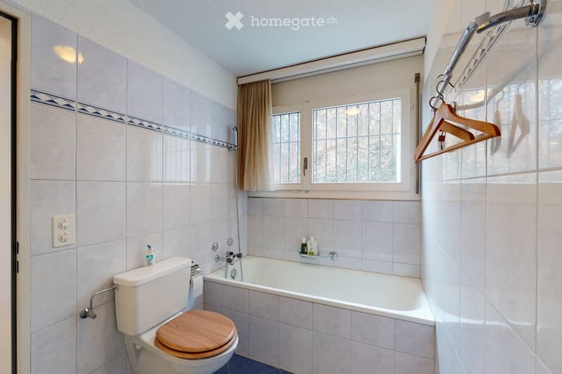 Bathroom with white tiled walls, a bathtub, toilet with wooden seat, and a window with a curtain. Wooden hangers are visible on the wall.