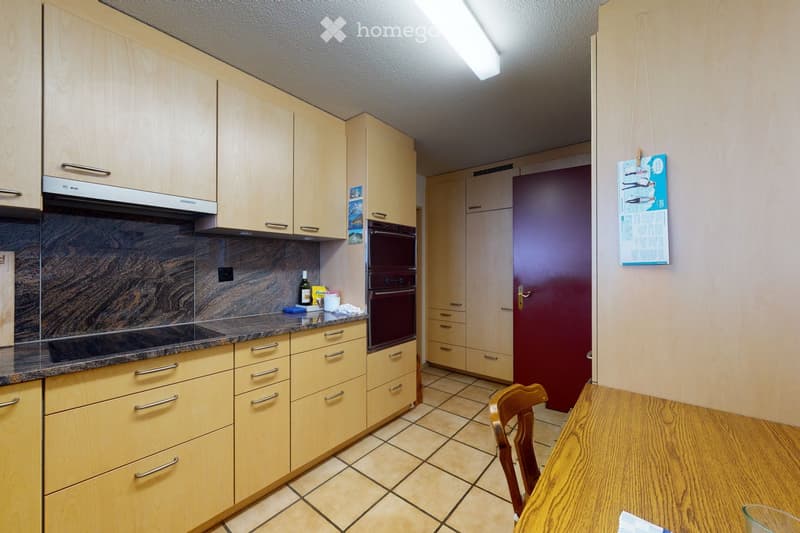 Bright kitchen with light wooden cabinets, granite countertop, and a small dining area with a wooden table and chair. A bottle and cup are placed on the counter.