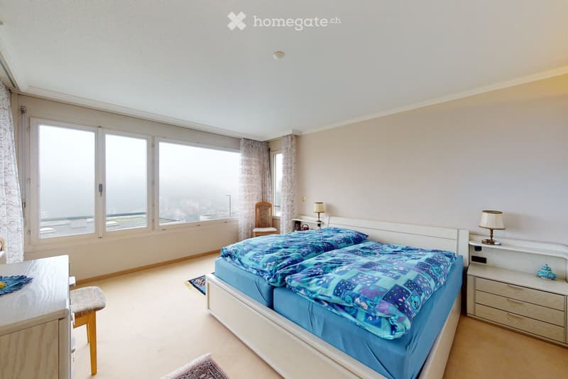 Bright bedroom with large windows, double bed with blue bedding, nightstands with lamps, a wooden chair, and a small table near the window.