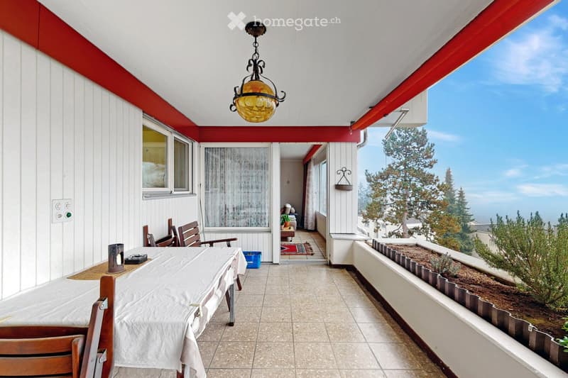 Balcony with a view, tiled floor, wooden chairs, table, hanging lamp, and small garden area.