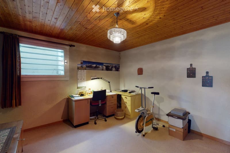 Cozy, well-lit room with wooden ceiling, large window, desk with chair, exercise bike, and storage boxes.