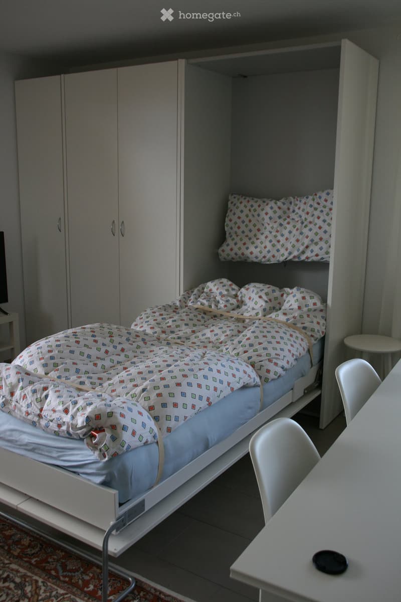 A compact bedroom with a single bed, a small table, and a chair, featuring built-in storage cabinets and a colorful bedspread.