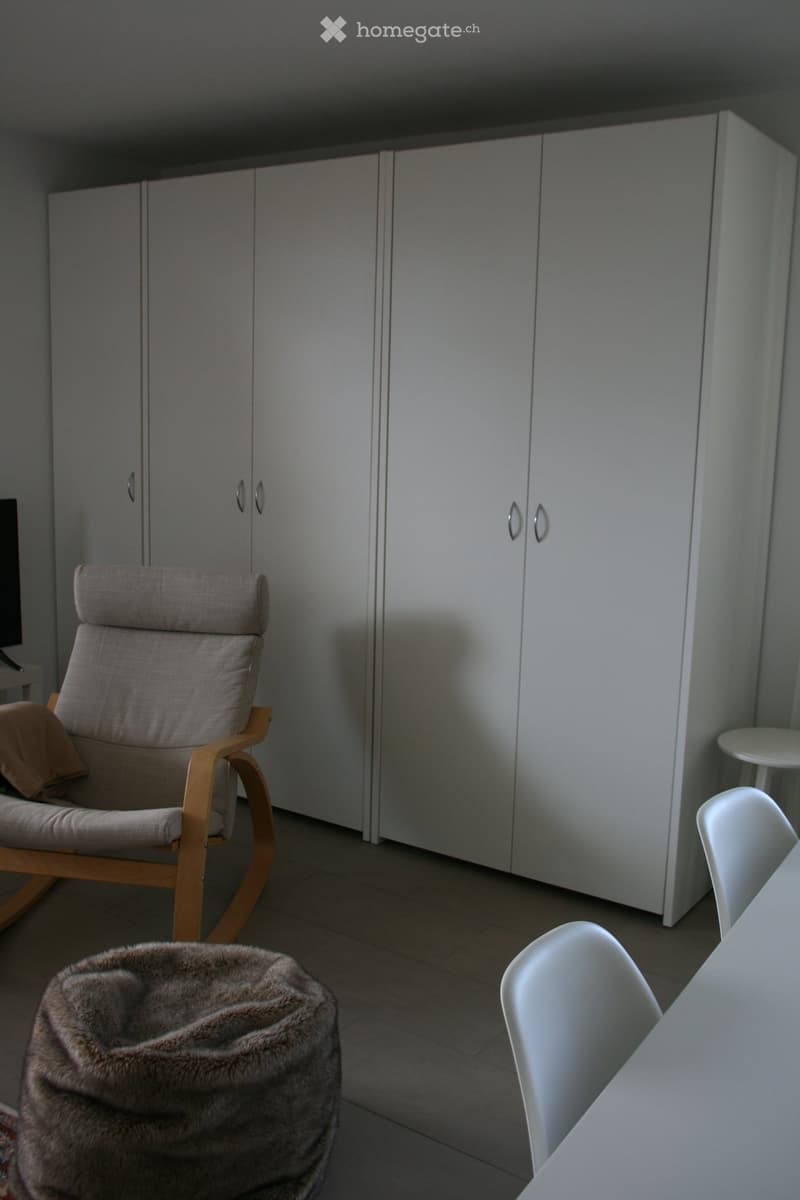 A modern, minimalist room with light-colored furniture, including a wooden chair, a small table, a white stool, and a furry rug on the floor.