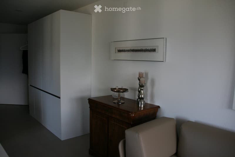 A small, modern room with a wooden table, a beige chair, a candle holder, and a picture frame on the wall. A white wardrobe is visible on the left side.