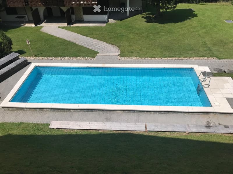 A rectangular swimming pool with blue tiles is surrounded by a paved area and grassy lawn, with a wooden deck and a small wooden bench nearby.