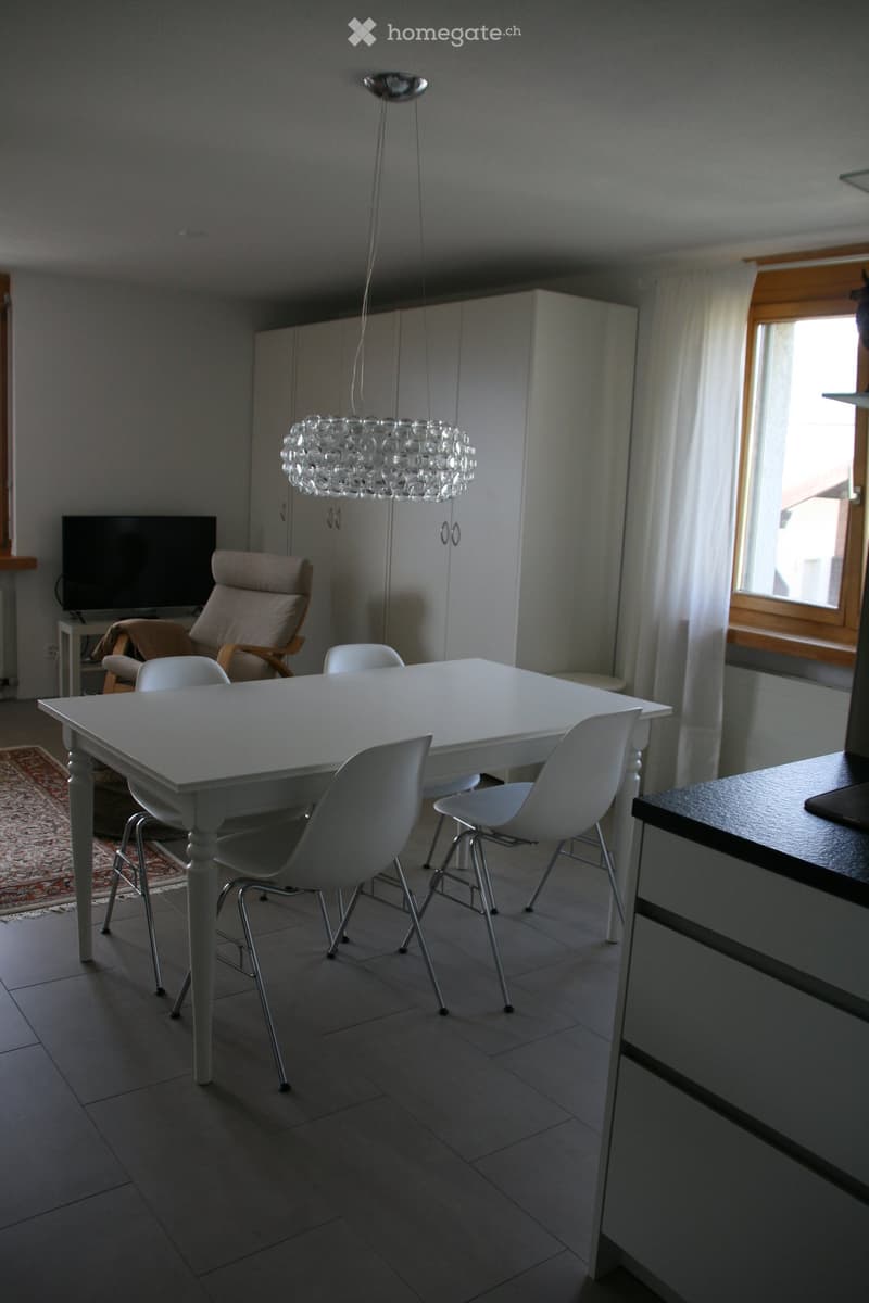 Dining room with a white table, four white chairs, a beige armchair, a modern chandelier, and a TV in the background.