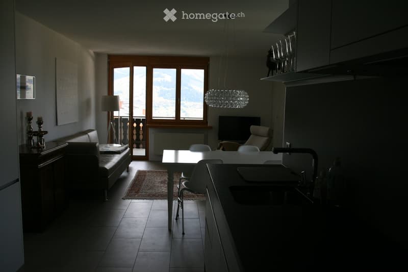 Open-plan living and dining area with a sofa, dining table, chairs, kitchen counter, sink, and television. Large windows offer a scenic mountain view.
