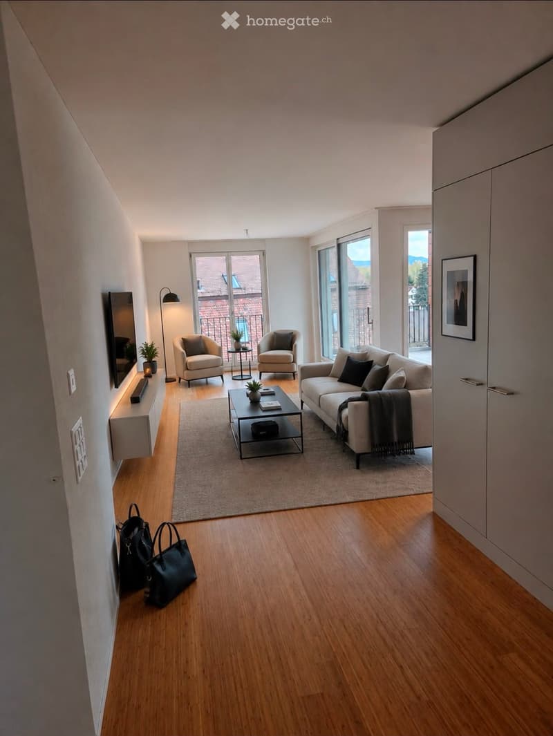 Modern living room with wooden flooring, large windows, balcony access, sofa, coffee table, armchairs, floor lamp, and mirror.