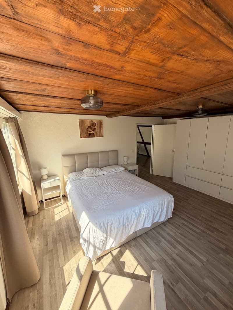 Cozy attic bedroom with wooden slanted ceilings, a large bed with white bedding, nightstands with lamps, a modern chair, and built-in white cabinets.
