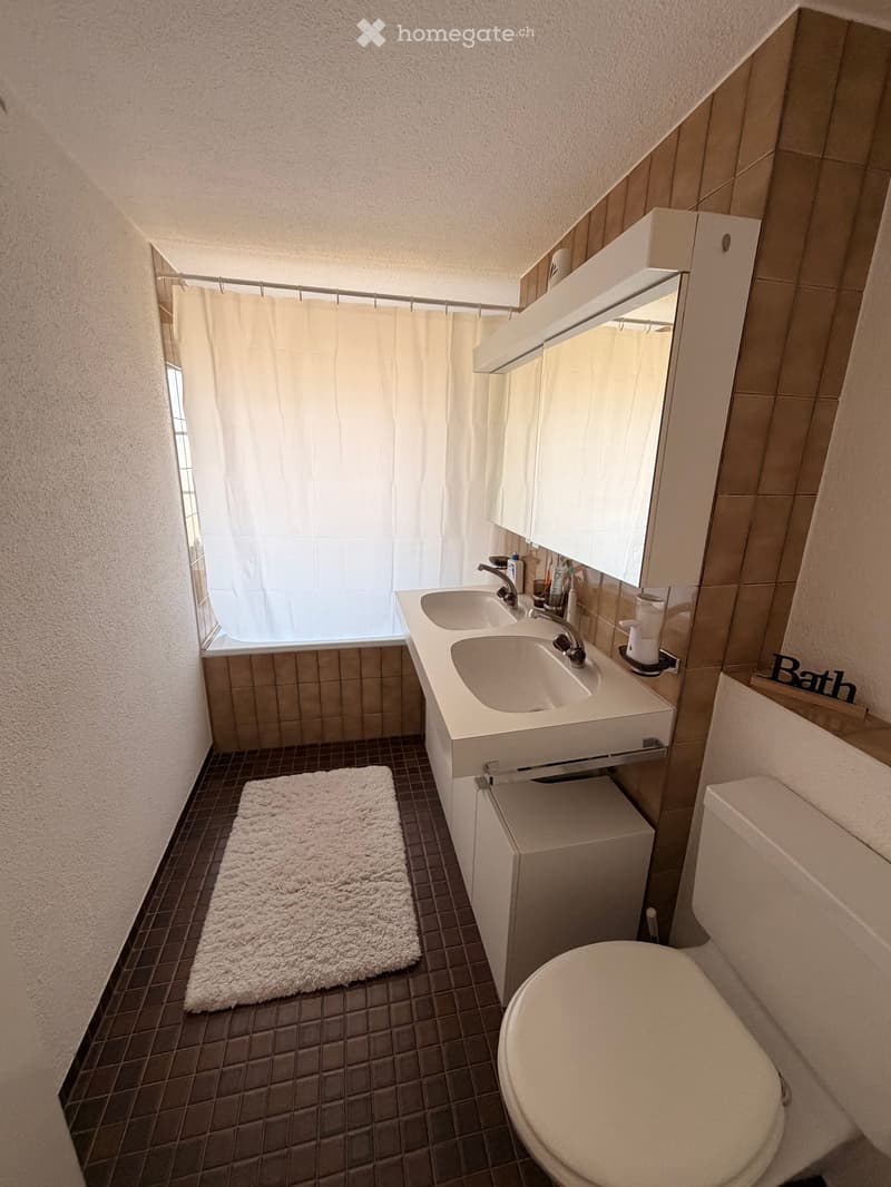 Modern bathroom with a double sink vanity, toilet, mirror, and a small shower area with a curtain. The floor has dark tiles, and a white fluffy rug is placed in front of the sink.