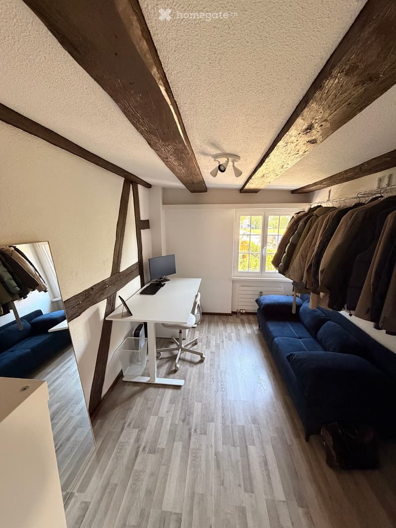 A modern, multi-functional room with exposed wooden beams, a blue sofa, a white desk with a computer, and a coat rack with jackets.