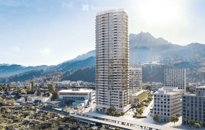 A modern high-rise building with a sleek design, surrounded by smaller structures and a scenic mountainous backdrop.