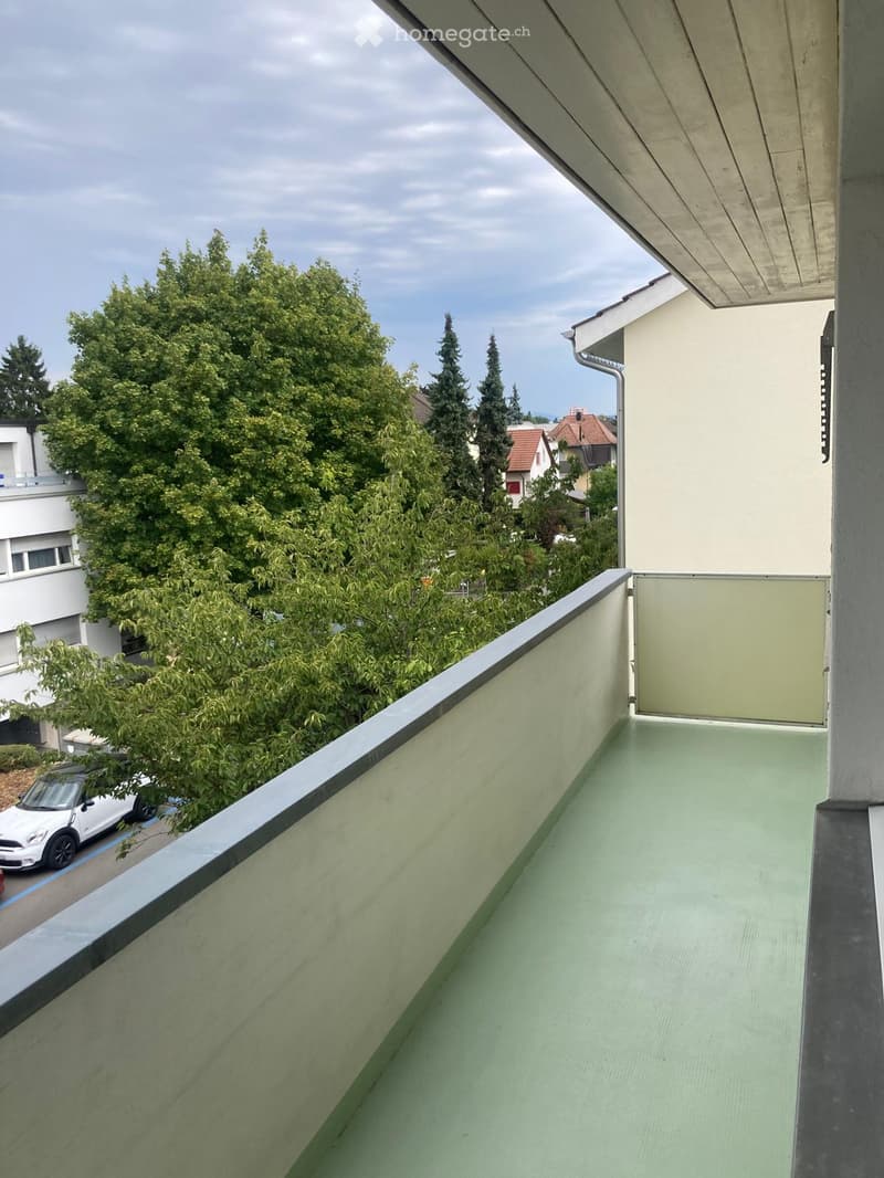 Balcony with green flooring, metal railing, and a view of trees and neighboring buildings.