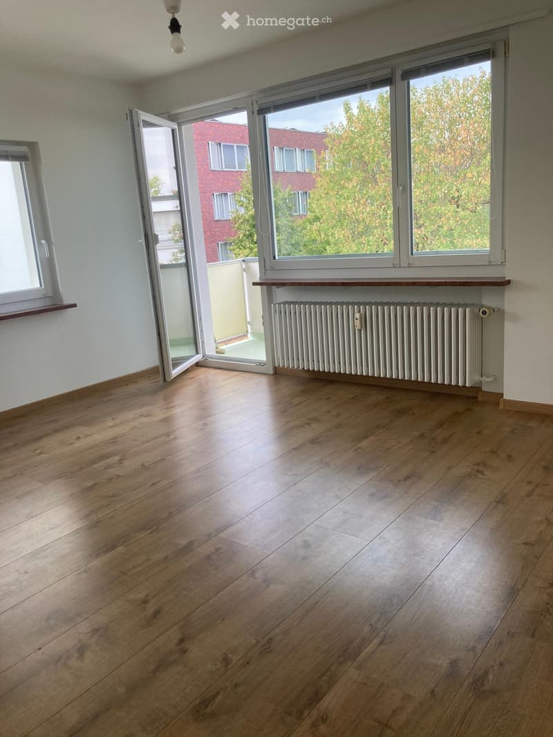 Empty room with wooden flooring, large windows, radiator, and open sliding doors leading to a balcony.