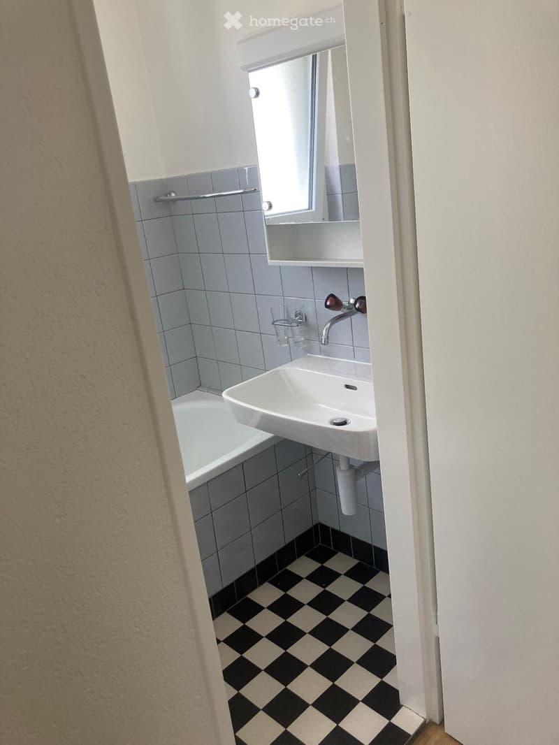 Small bathroom with a bathtub, white sink, mirror, and tiled walls. The floor features a black and white checkered pattern.