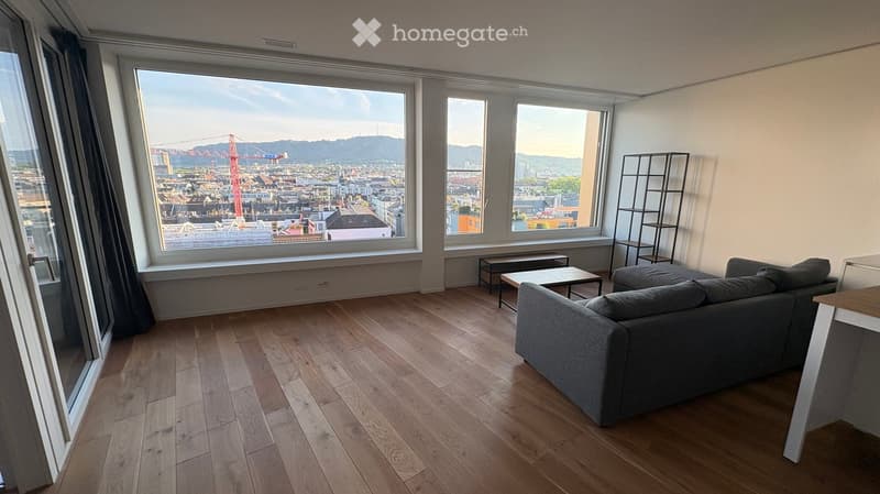 Modern living room with wooden flooring, large windows offering a cityscape view, a grey sofa, a coffee table, and a tall shelving unit.