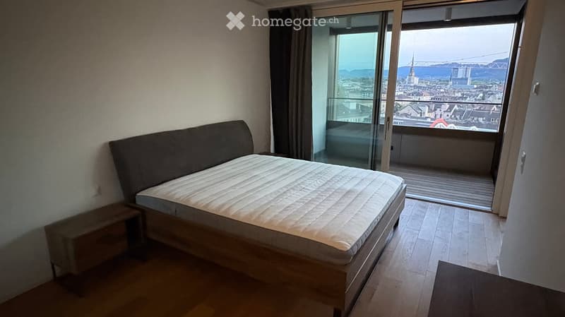 Modern bedroom with a single bed, nightstand, and large windows offering a cityscape view.