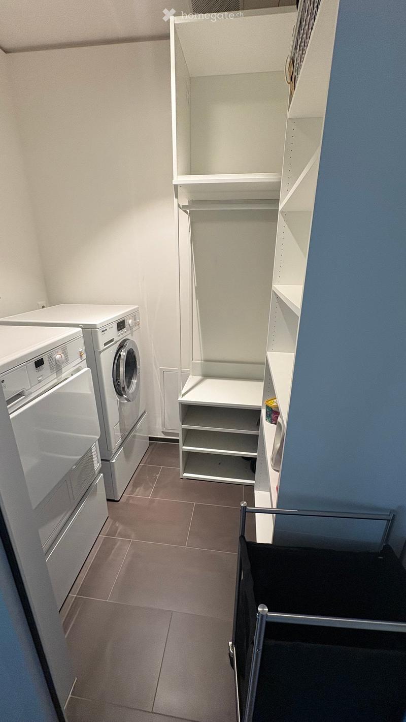 A small, well-lit laundry room with a washing machine, dishwasher, and built-in white shelves. The floor is tiled, and the walls are painted in neutral tones with a blue accent wall.