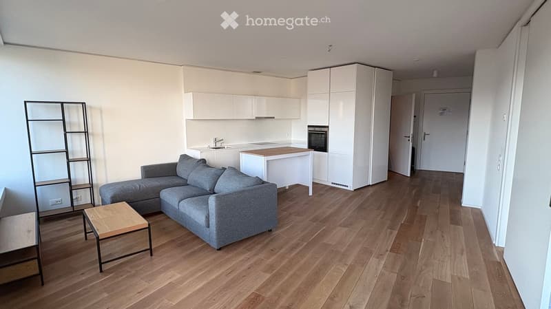 Modern living and dining area with a grey sectional sofa, wooden coffee table, kitchen island, white cabinets, sink, and oven. The space features light wooden flooring and white walls.