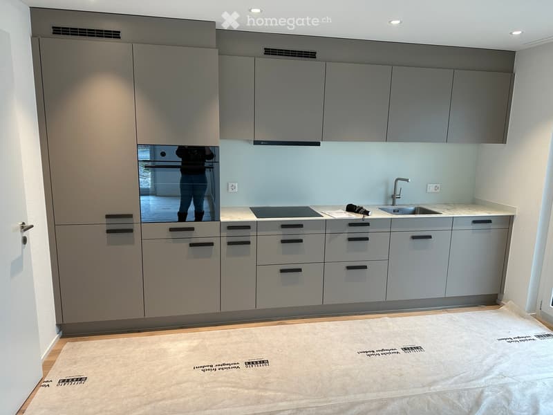 Modern kitchen with grey cabinets, stainless steel oven, sink, and countertop. Light blue backsplash and wooden flooring.