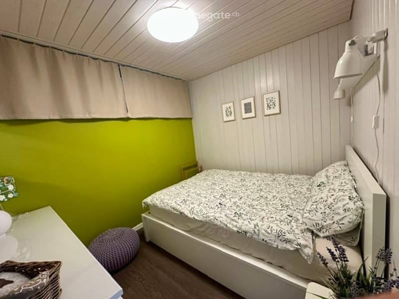 Cozy bedroom with a single bed, nightstand, small stool, and wall-mounted lamps. The walls are painted in a light green and white color scheme, with framed pictures on the wall.