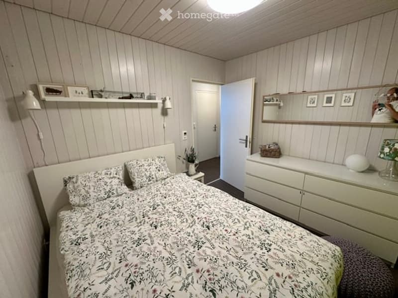 Cozy bedroom with wooden panel walls, a bed with floral bedding, a nightstand, a mirror, a drawer unit, and a shelf with decorative items.